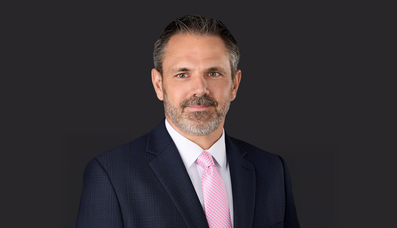 Bearded professional man in a navy suit with a light pink tie against a dark background