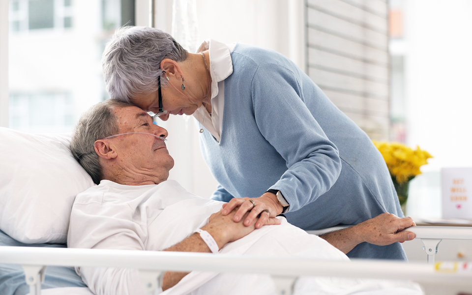 Woman gently leaning over and holding hands with a man lying in a hospital bed.