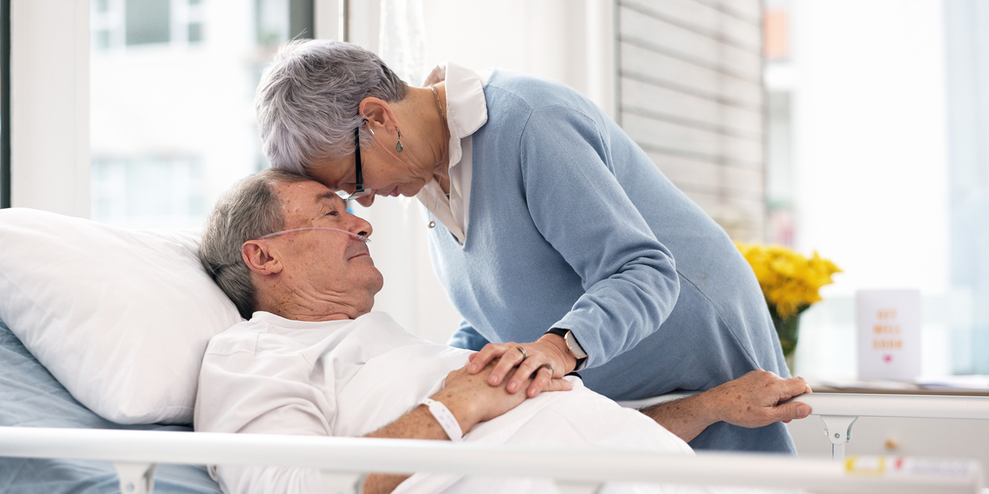 Woman gently leaning over and holding hands with a man lying in a hospital bed.