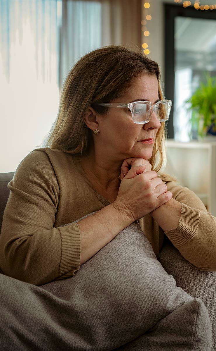 Thoughtful middle-aged woman sitting indoors, reflecting quietly with hands clasped on a pillow