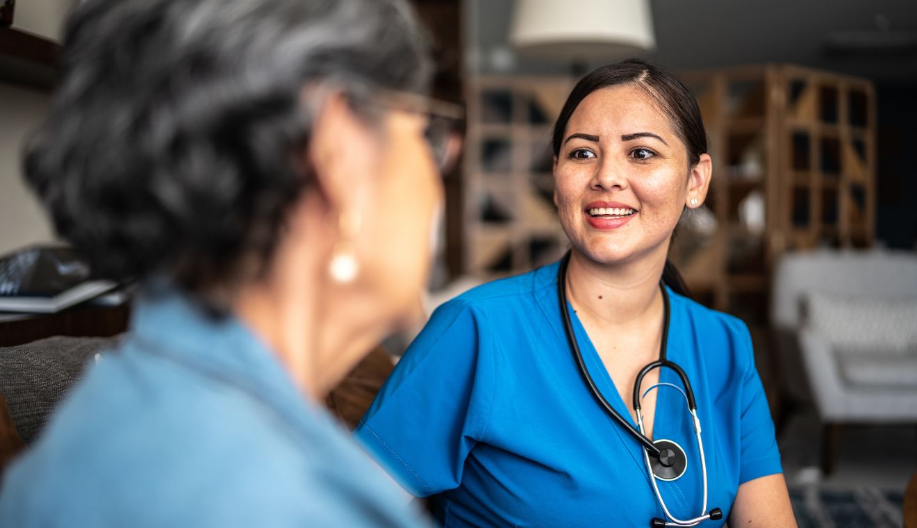 Healthcare professional in blue scrubs speaking with and attentively listening to an older adult during a compassionate in-home care visit or patient consultation.